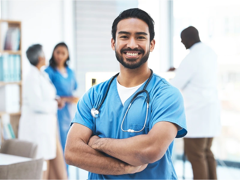 A healthcare professional in blue scrubs stands confidently with arms crossed. Three colleagues in medical attire are conversing in the blurred background of a bright, modern medical facility.