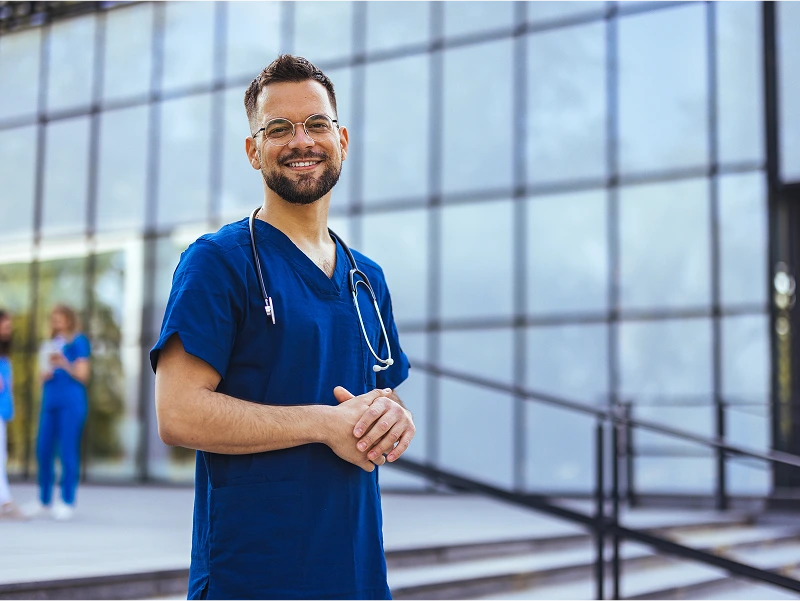 A man in blue scrubs, wearing glasses and a stethoscope, smiles in front of a modern glass building. Two people in scrubs are visible in the background.