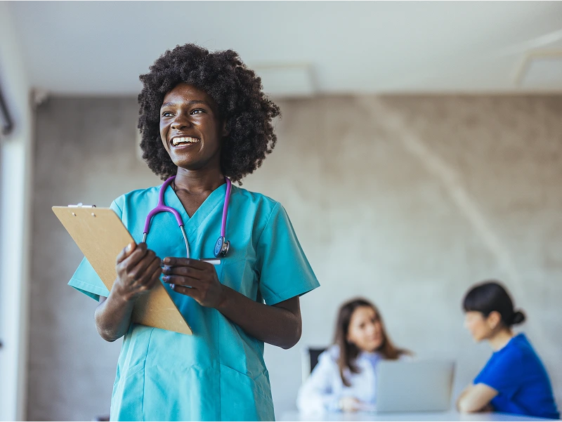 A smiling healthcare professional in green scrubs holds a clipboard. Two colleagues are sitting and working at a table in the background.