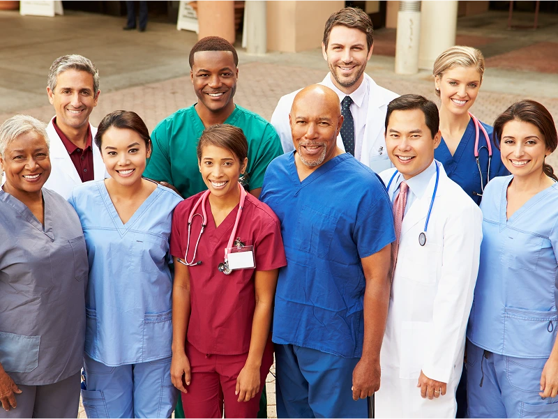 A group of medical professionals, including doctors and nurses, are standing together and smiling. They are wearing scrubs, lab coats, and stethoscopes.